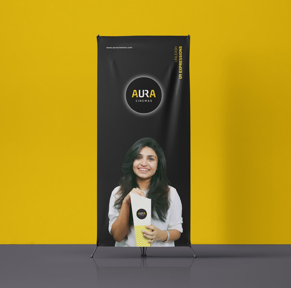 Aura Cinemas promotional roll-up banner in Bengaluru featuring the logo, a smiling woman holding popcorn, and the slogan 'UNLEASH UR EXPRESSIONS' on a black background.