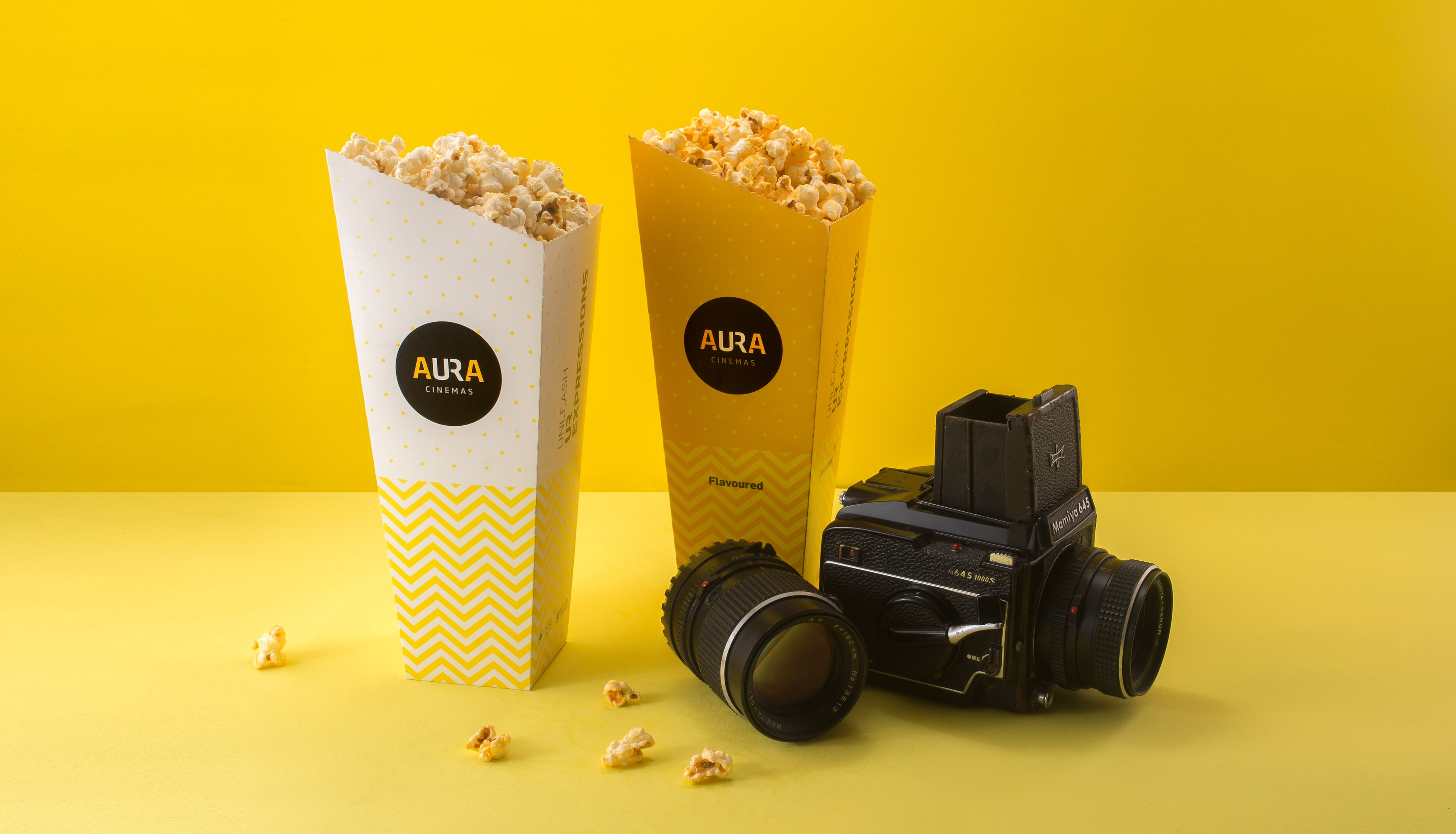 Aura Cinemas branded flavored popcorn in yellow zigzag-patterned containers alongside a vintage camera, enhancing the cinematic snack experience