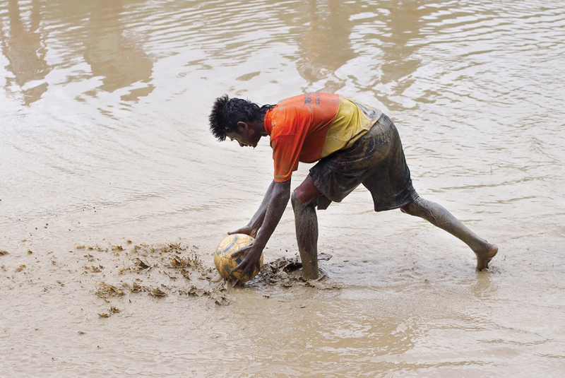 A male soccer player, covered in mud, bends down to retrieve a muddy ball from a waterlogged field during a monsoon carnival, representing an event like Splash.